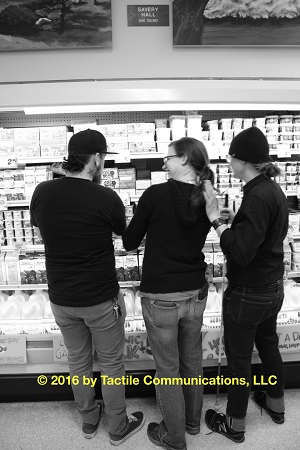 Image description: b&w picture of three people standing in front of a dairy section in Trader Joe’s. There are stacks of different types of dairy products on the shelves in the background; a male, Ray, signs with Hayley. aj has her left hand on Hayley’s shoulder, while her right hand holds a white cane. *Image © 2016 by Tactile Communications, LLC