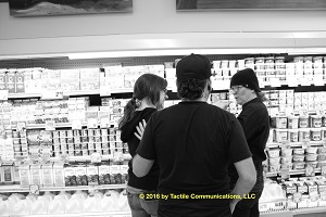 Image description: b&w picture of three people, two females and one male, standing in front of a dairy section in Trader Joe’s.Hayley, wearing a pair of glasses, is standing to the left, Ray with his back to the camera is standing in the middle, and aj is standing to the right. aj also has a pair of glasses on. While aj is tactiling with Hayley, Ray looks on with his left hand on Hayley’s shoulder. *Image © 2016 by Tactile Communications, LLC