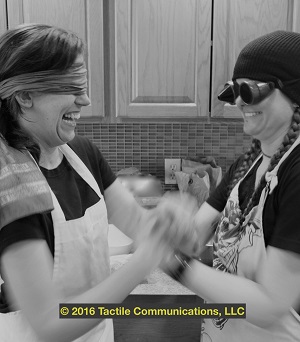 Image description: black and white image of two women (Hayley and aj) standing in front of a kitchen counter, one blindfolded and the other has a pair of goggles on, ProTactiling. The woman blindfolded is laughing, while the woman wearing a pair of goggles is smiling big. *Image © 2016 Tactile Communications, LLC
