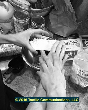 Image description: black and white image of a pair of hands and a hand; a hand is resting over one pair of hands (ProTactiling). At the same time, these pair of hands are holding a measuring cup over a bowl, which is filled with almond flour. In the background, there are different items on the counter. *Image © 2016 Tactile Communications, LLC