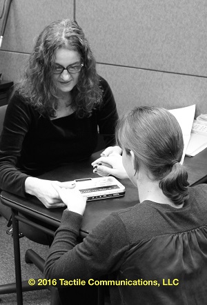 Image description: b&w picture of two women, Jelica and CJ. Both are at a small table; CJ’s back is to the camera, while Jelica looking down, away from the camera. Jelica is showing CJ the braille notetaker; a device called, BSU2. *Image © 2016 by Tactile Communications, LLC