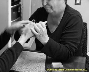 Image description: b&w picture of a woman, Theresa, whom is wearing a black long sleeve top, using PT with another person whose face is not seen except for their hands (their nails are painted black). Theresa, pictured, is "drawing" a map on the person's hand.  *Image © 2016 by Tactile Communications, LLC