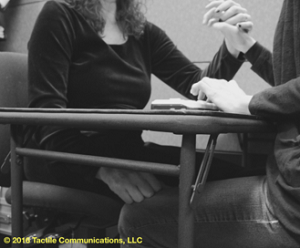 Image description: b&w picture of two people sitting at a table with a braille device, both wearing a black long sleeve shirt, and both are ProTactiling. The woman on the left, has her left hand on the person opposite of her, while her right hand is under the table, tapping (yes) on the person's knee. *Image © 2016 by Tactile Communications, LLC